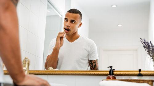 Young man brushing teeth reflecting on mirror in bathroom