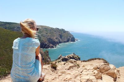 Rear view of woman sitting on rock by sea against clear sky