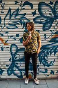 Full body of trendy young male with long hair holding cold ice cream in one hand and keeping another in pocket while standing against painted wall and looking away on summer evening