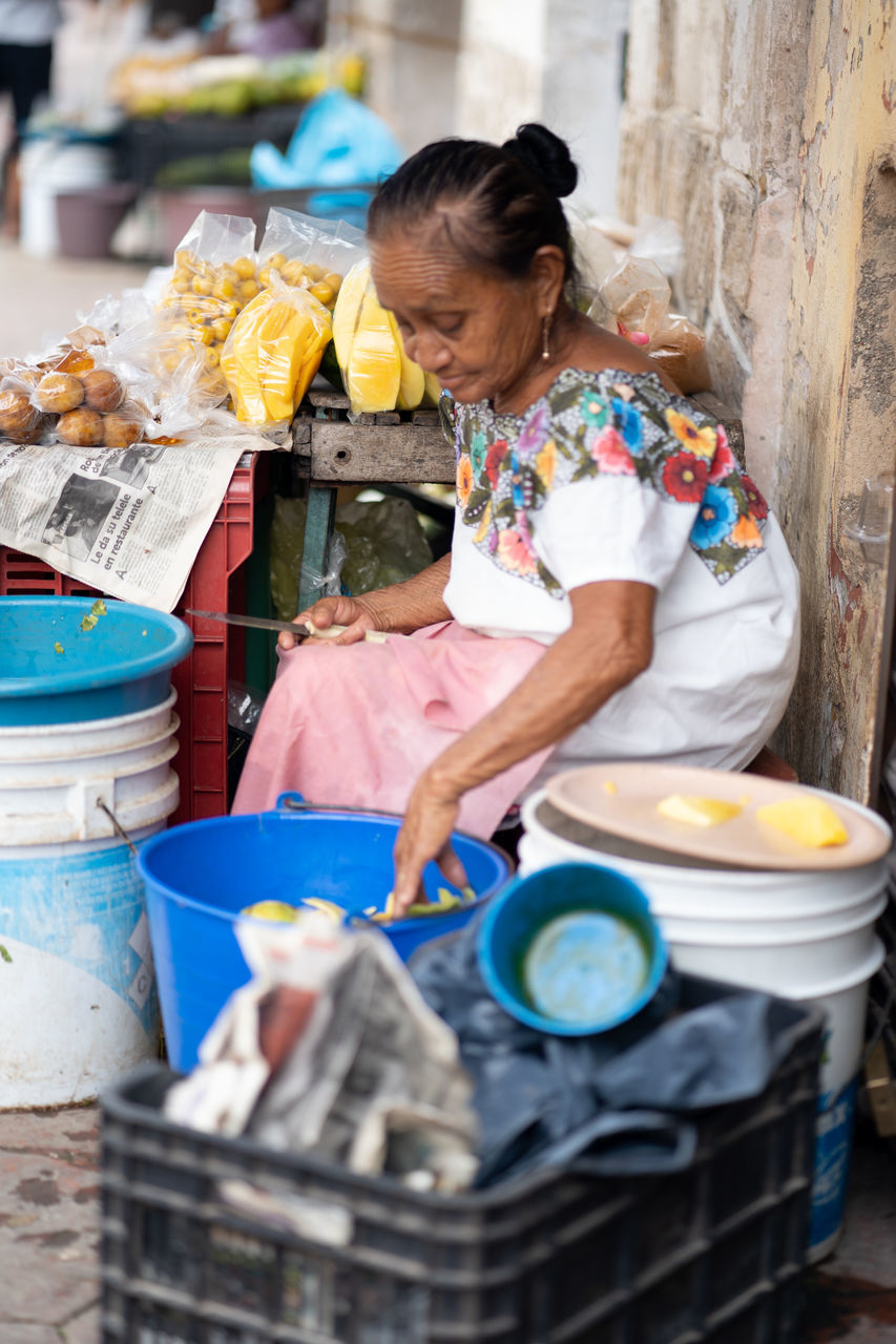 adult, one person, women, market, business, occupation, business finance and industry, small business, food and drink, food, retail, working, selling, vendor, female, market stall, child, container, mature adult, holding, standing, public space, street market, city, looking down, person, outdoors