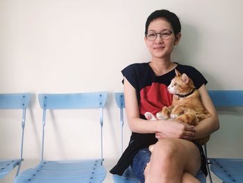 Smiling woman with cat looking away sitting on chair against wall