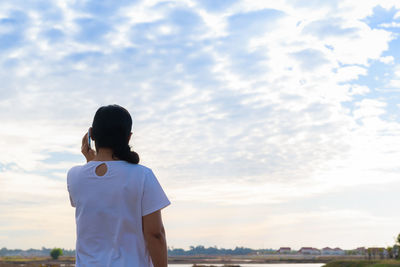 Rear view of woman answering smart phone while standing against cloudy sky