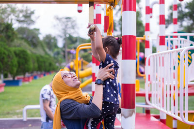 Mother assisting daughter in hanging on monkey bars at playground