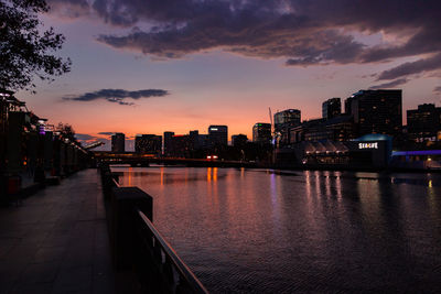 Illuminated buildings by river against sky during sunset