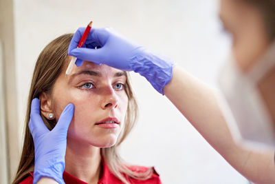 Blur beautician in latex gloves drawing line with white pencil while correcting shape of eyebrows of young woman during work in aesthetic clinic