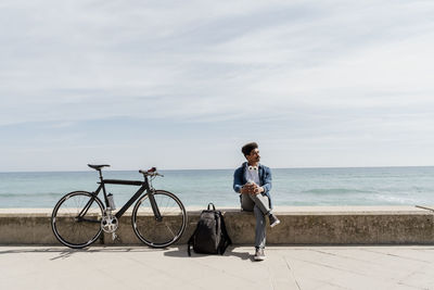 Man with legs crossed at knee sitting on retaining wall by backpack during sunny day