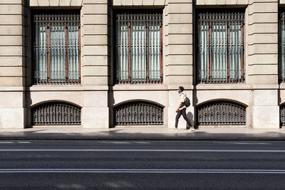 Side view of black male in medical mask walking along pavement near urban building on sunny day in barcelona during coronavirus epidemic