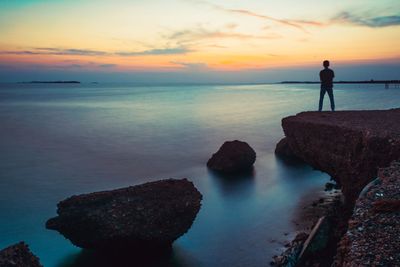 Rear view of silhouette man standing on cliff by sea against dramatic sky during sunset