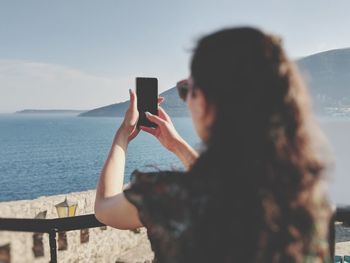 Rear view of woman taking selfie while standing against sea and sky