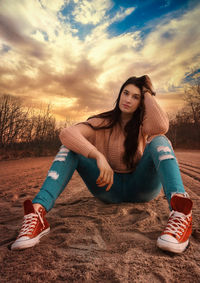 Beautiful young woman sitting on field against sky during sunset