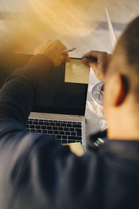 High angle view of man writing on adhesive note over laptop at table