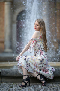 Side view of beautiful young woman sitting against fountain in city