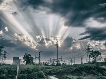 Electricity pylon by factory on field against sunbeams emitting from cloudy sky