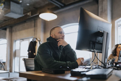 Young male hacker with hand on chin using computer at desk in creative office