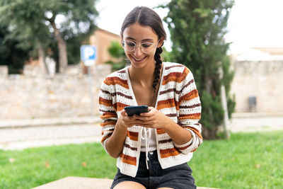 Positive young female sitting on bench in city and messaging on social media via mobile phone while looking at screen