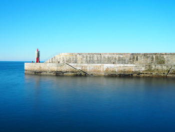 Lighthouse on pier by seascape against clear blue sky