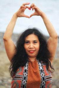 Portrait of smiling mature woman with arms raised making heart shape while standing against sky