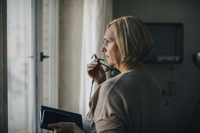 Mature businesswoman talking through earphones while standing by window in creative office