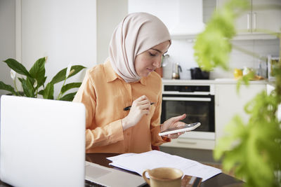 Smiling woman with hijab using cell phone during work from home in kitchen