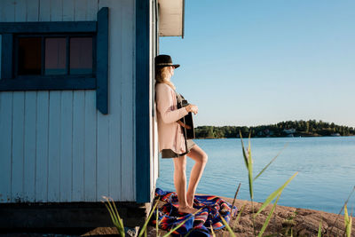 Woman leaning against a wooden beach hut playing the guitar in the sun