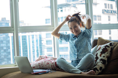 Young woman using mobile phone while sitting on window