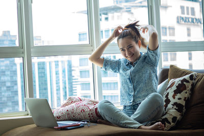 Young woman using mobile phone while sitting on window