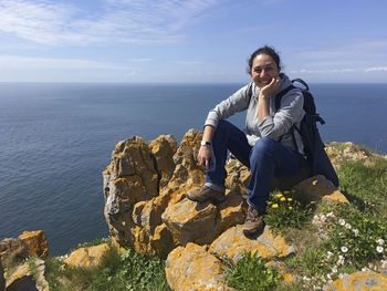 Portrait of female hiker smiling while sitting on cliff against sea