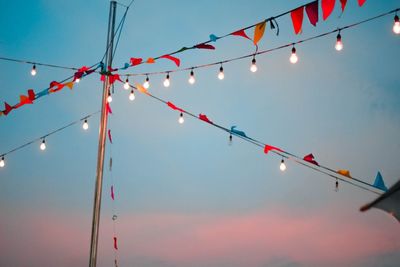 Low angle view of bunting flags and light bulbs hanging against sky during sunset