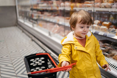 Child in the market with a grocery cart, wearing a yellow jacket and jeans