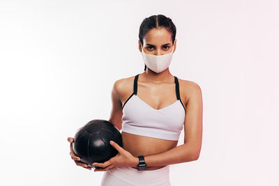 Portrait of young woman wearing mask with medicine ball against white background
