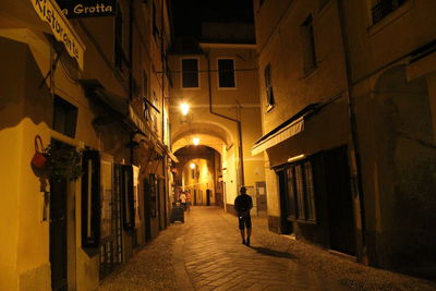 Rear view of man walking on narrow street amidst buildings at night