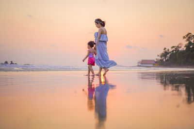 Mother with daughter walking at beach against sky during sunset