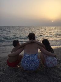 Rear view of couple sitting on beach against sky during sunset