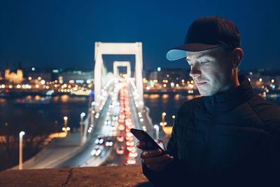 Young man using phone against illuminated bridge in city at night