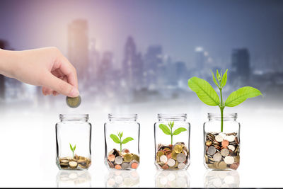 Cropped hand putting coin in jar with seedling on table against city at night