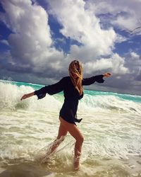 Young woman enjoying amidst waves in sea against cloudy sky