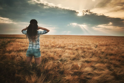 Rear view of woman standing amidst plants on field against sky during sunset