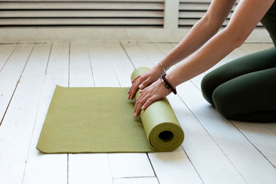 Young slender woman's hands twisting green mat for fitness or yoga on white wooden floor.