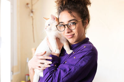 Happy young curly haired hispanic female owner in eyeglasses cuddling cute domestic cat and looking at camera while resting together at home