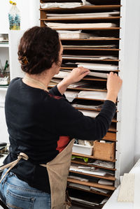 Back view of craftswoman in apron taking paper sheet from shelf while preparing material for printmaking in workshop