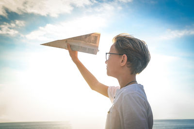Boy holding paper airplane standing by sea against sky