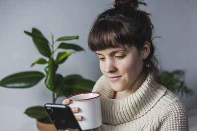 Peaceful female in warm sweater and with mug of hot drink messaging on smartphone during weekend at home