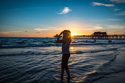Girl standing on shore at beach against sky during sunset