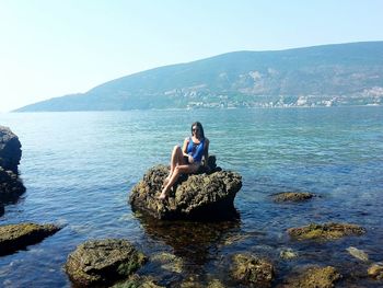 Woman sitting on cliff by sea against clear sky