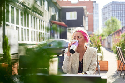 Low angle of pensive alternative female with dyed hair drinking hot beverage from eco friendly cup while sitting at table in street cafe and looking away