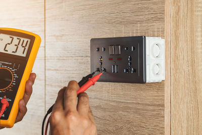 Electrician using a digital meter to measure the voltage at a wall socket on a wooden wall.