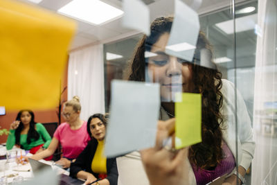 Young female entrepreneur writing on adhesive note during business meeting with colleagues at office