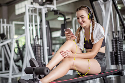 Young woman listening music over mobile phone while sitting on equipment in gym