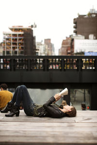 Full length of woman reading book while lying on boardwalk in city