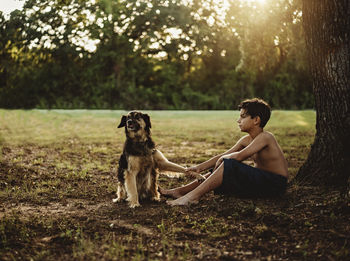 Side view of shirtless teenage boy sitting with dog on field